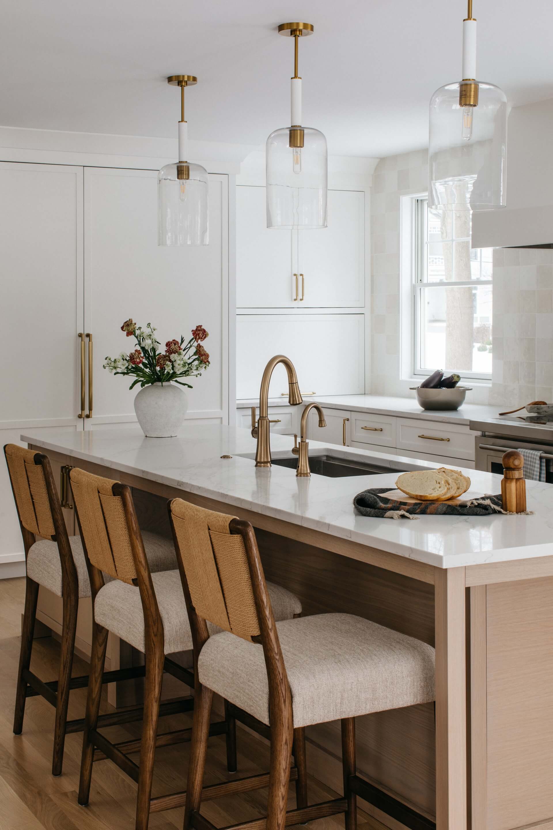 Remodeled white kitchen features a marble island, brass fixtures, woven chairs, pendant lights, and fresh flowers.